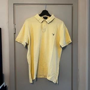 Ljusgul pikétröja från GANT - Säljer en ljusgul pikétröja från GANT i Regular Fit. Klassisk krage och korta ärmar, perfekt för en stilren och avslappnad look. Diskret logga broderad på bröstet. Passar dig som gillar tidlös stil.
