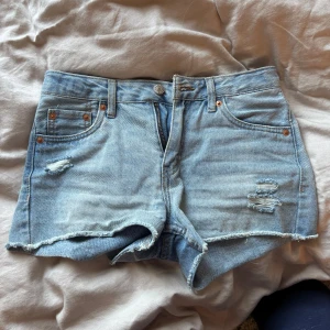Ljusblå jeansshorts från Levi's - Säljer ett par ljusblå jeansshorts från Levi's med slitna detaljer och råa kanter. Klassisk femficksmodell med knapp och dragkedja framtill. Perfekta för varma dagar och enkel att matcha med allt.