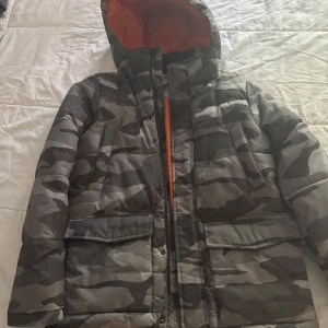 Camouflage pufferjacka från McKinley - Säljer en camomönstrad pufferjacka från McKinley med huva och orange innerfoder. Jackan har dragkedja, stora fickor framtill och är perfekt för kyligare dagar. Snygg och praktisk jacka för dig som gillar streetstil.