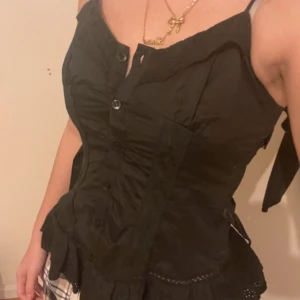 Svart korsett-topp med spetsdetaljer - Säljer en svart korsett-topp med knappar framtill och fina spetsdetaljer längs nederkanten. Toppen har en figurnära passform och volang längst ner för en extra touch.