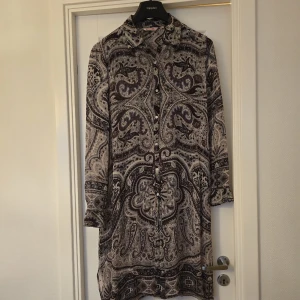 Mönstrad skjortklänning från Indiska - Säljer en långärmad skjortklänning från Indiska i storlek L. Klänningen har ett detaljerat paisleymönster i mörka och ljusa toner, knappar framtill och klassisk krage. Perfekt för dig som gillar bohemiska och unika plagg.
