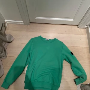 Grön sweatshirt från Stone Island Junior - Snygg grön sweatshirt från Stone Island Junior med rund halsringning och klassisk logotyp-patch på vänster ärm. Tröjan har långa ärmar och ribbade muddar vid ärmslut och nederkant. Perfekt för en avslappnad stil.