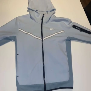 Ljusblå hoodiejacka från Nike - Nike hoodiejacka i ljusblått med vit logga på bröstet och vita detaljer längs dragkedjan. Jackan har huva, hel dragkedja och lång ärm. Perfekt för en sportig look. nyskick bara aty färgen inte är samma som ifall den va nyköpt. Storlek S funkar till M.