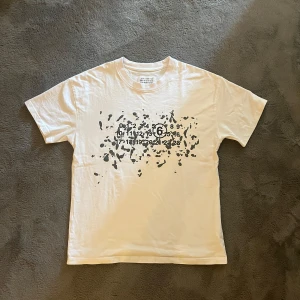 Vit t-shirt från Maison Margiela - Vit t-shirt från Maison Margiela med svart grafiskt tryck och ikoniska siffror på bröstet. Klassisk passform och rund halsringning. Tillverkad i mjuk bomull och made in Italy.   Nypris: €390 (cirka 4 300 kr) 	•	Mitt pris: 599 kr 	•	Skick: Mycket bra, sparsamt använd 	•	Storlek: S/M