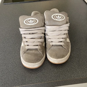 Gråa Adidas sneakers med snörning - Säljer ett par gråa Adidas sneakers med klassisk vit logga på plösen och baksidan. Skorna har snörning och är tillverkade i mocka med vit sula. Perfekta för en avslappnad och sportig stil. Kommer med 2 extra snören gråa 