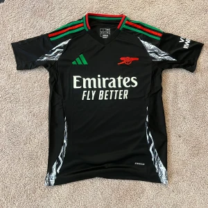 Arsenal borta svart fotbollströja från Adidas - Säljer en svart Arsenal bortatröja från Adidas med gröna och röda detaljer på axlarna. Tröjan har klubbens kanon-logga i rött, Emirates Fly Better-tryck på bröstet och mönstrade inslag i vitt och grått på sidorna. Klassisk rund hals och korta ärmar.