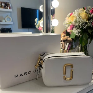 Vit axelväska från Marc Jacobs - Säljer en snygg vit axelväska från Marc Jacobs med guldiga metalldetaljer och bred axelrem med logga. Väskan har två dragkedjor och stilren design, perfekt för att lyfta vilken outfit som helst.