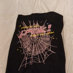 Svart hoodie med rosa spindelnäts-tryck - Svart hoodie med stor rosa spindelnätsgrafik och texten 'Pink!' i rosa nitar framtill. Gula och blå stjärnor runt trycket. Huva och klassisk passform. Perfekt för dig som gillar streetwear med attityd. Tar bara kontant möts upp i Eskilstuna Örebro Arboga 