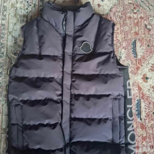 Svart dunväst från Moncler - Snygg svart dunväst från Moncler med hög krage och diskret logga på bröstet. Västen har breda vadderade paneler, dragkedja framtill och stora Moncler-tryck längs sidorna. Perfekt för lager-på-lager och streetstyle. Finns även QR kod som går direkt till hemsidan 