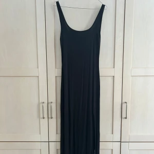 Svart långklänning med slits Ginatricot - Säljer en svart långklänning med djup urringning och slits framtill. Klänningen är ärmlös och har en tight passform som framhäver figuren ovantill, och lösare vid benen. Har en djupare ringning i ryggen