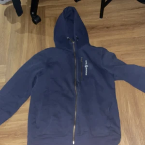 Blå hoodie från Sail Racing - Mörkblå hoodie från Sail Racing med dragkedja framtill och logga på bröstet. Tröjan har huva och en ficka med dragkedja på bröstet. Perfekt för dig som gillar sportig stil.