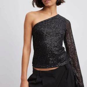 Svart paljetttopp från NA-KD - Säljer en svart enarmad topp från NA-KD täckt med glittriga paljetter. Perfekt för dig som vill sticka ut med en elegant och festlig. 