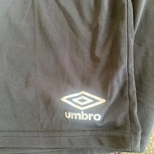 Svarta träningsshorts från Umbro - Säljer ett par svarta träningsshorts från Umbro med klassisk logga framtill. Shortsen har elastisk midja och är tillverkade i ett lätt och svalt material som passar perfekt för sport och träning.