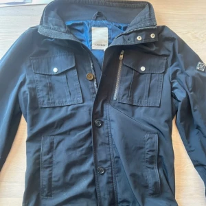 J.Lindeberg Field Jacket Navy - Stilren Field Jacket från J.Lindeberg i mörkblått, storlek M, i mycket fint skick. En mångsidig jacka med både funktion och design – passar utmärkt till både jeans och kostymbyxor. Perfekt jacka för övergångsperioder.