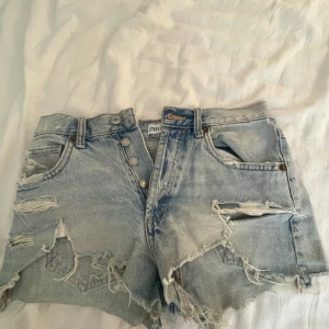 Ljusblå slitna jeansshorts från ZARA - Ljusblå jeansshorts från ZARA med medelhög midja. Shortsen har knappar framtill, klassiska fickor och tydliga slitningar för en edgy look. Perfekta för sommaren och festivaler. Mycket använda därav priset, zooma in och titta på bild 5 för defekter i detalj! 