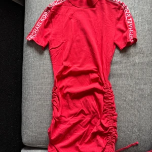 Korallfärgad kortklänning från Michael Kors - Säljer en korallfärgad kortklänning från Michael Kors med hög krage och korta ärmar. Klänningen har logotypband längs axlarna och en snygg rynkdetalj på sidan som kan justeras med snörning. Orginpris ca 2500kr
