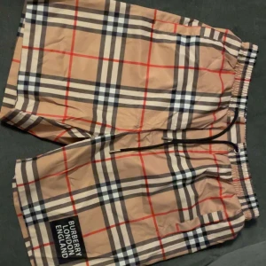 Beige rutiga shorts från Burberry - Säljer ett par klassiska beige shorts från Burberry med svart, vitt och rött rutmönster. Shortsen har resår i midjan med svart snörning och en Burberry London England-patch på benet. Perfekta för sommaren!