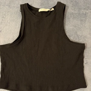 Svart ribbad croppad linnetopp från Black Story - Säljer en svart ribbad croppad linnetopp från Black Story i storlek XS. Perfekt till sommaren och enkel att matcha med olika outfits. Snygg och stilren design med ärmlöst snitt.