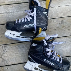 Svarta hockeyskridskor från Bauer - Säljer ett par svarta hockeyskridskor från Bauer med vita snören och detaljer i gult och vitt. Skridskorna har en robust konstruktion och klassisk design, perfekta för isträning eller match. Sulan och hälpartiet är förstärkta för extra stabilitet. Bauer Supreme s150 i storlek 36