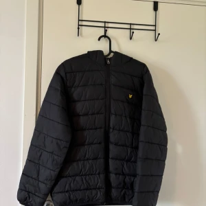 Svart pufferjacka från Lyle & Scott Junior - Svart pufferjacka från Lyle & Scott Junior med huva och dragkedja framtill. Jackan har quiltad design och en liten gul logga på bröstet. Perfekt för kyligare dagar och enkel att matcha med olika outfits.
