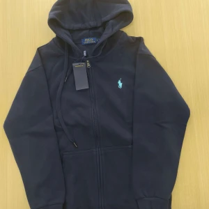 Mörkblå hoodie från Polo Ralph Lauren - Säljer en mörkblå hoodie från Polo Ralph Lauren med dragkedja och huva. Tröjan har en broderad logga i ljusblått på bröstet och två fickor framtill. Perfekt för en avslappnad stil.