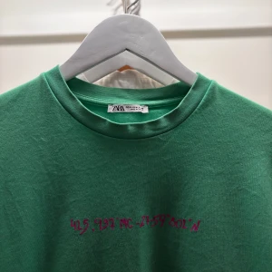 Grön t-shirt från Zara med tryck - Säljer en grön t-shirt från Zara med rosa texttryck på bröstet. T-shirten har rund halsringning och korta ärmar. Perfekt för en avslappnad stil.