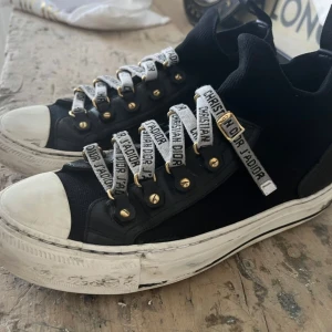 Svarta sneakers från Christian Dior - Snygga svarta sneakers från Christian Dior med vita snören där det står 'Christian Dior J'Adior'. Skorna har vita sulor med svart stjärndetalj undertill och guldfärgade öljetter. Tydlig branding baktill på sulan. Säljer då dem är för små för mig. Är i använt skick och sulan har defekt på båda skorna därav priset, och rekommenderar att man använder dem varsamt pga detta. Annars är skon i övrigt i fint skick. Säljes med extra skosnören & äkthetsbevis Nypris är 10 500 kr. 