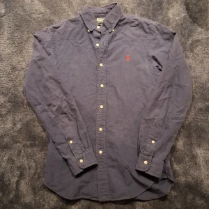 Mörkblå skjorta från Ralph Lauren - Klassisk mörkblå skjorta från Ralph Lauren med röd broderad logga på bröstet. Skjortan har knappar hela vägen och button-down krage. Perfekt för en stilren look.