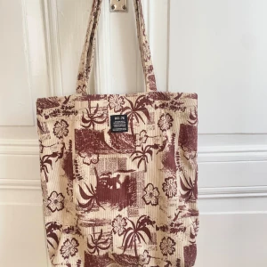 Väska från Urban Outfitters - Säljer en shoppingväska med vinrött tropiskt mönster från Urban Outfitters. Väskan har långa handtag och är perfekt för att bära med sig till stan eller skolan. Rymlig.