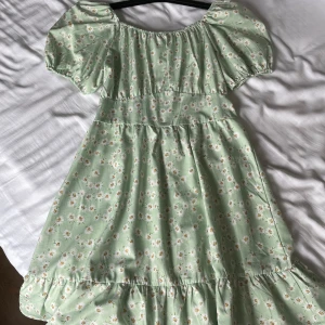 Ljusgrön blommig klänning med puffärmar - Superfin ljusgrön klänning med små vita och gula blommor. Klänningen har puffiga kortärmar, resår vid halsringningen och en söt volang längst ner. Dragkedja i ryggen för enkel påklädning. Perfekt för sommarens alla tillfällen!