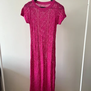 Stickad rosa långklänning - Säljer en stickad långklänning i stark rosa färg med korta ärmar och rund hals. Klänningen har ett detaljerat mönster i stickningen som ger en unik look. Perfekt för dig som vill sticka ut med färg och stil.