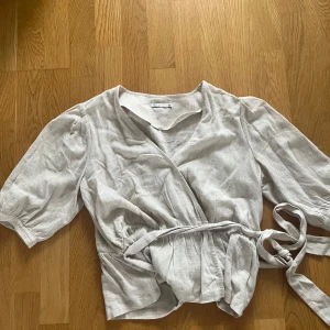 Beige omlottblus med knyt från NLY Trend - Säljer en beige omlottblus från NLY Trend i storlek 40. Blusen har korta puffärmar och knytband i midjan för en snygg passform. Tillverkad i ett linneliknande material som känns luftigt och skönt.