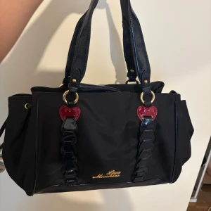 Svart handväska från Love Moschino - Säljer en svart handväska från Love Moschino med två handtag och dekorativa röda hjärtan framtill. Väskan har guldfärgade detaljer och flera fack inuti. Perfekt för dig som vill ha en snygg och rymlig accessoar.