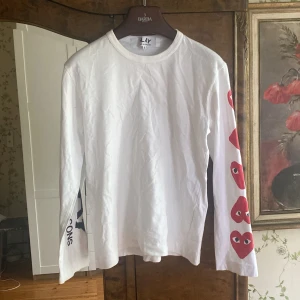 Comme des Garçons tröja  - Vit långärmad tröja från Comme des Garçons Play med röda hjärtan längs ena ärmen och svart text på den andra. Rund halsringning och avslappnad passform. Perfekt statement-plagg för dig som gillar streetwear. Storlek S passar XS