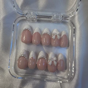 Press-on naglar med pärlor och rosetter - Säljer ett set med press-on naglar i ljusrosa och vitt med fransk tipp. Dekorerade med små pärlor och 3D-rosetter för en söt och elegant look. Kommer i en genomskinlig ask, perfekta för att snabbt fixa snygga naglar hemma.