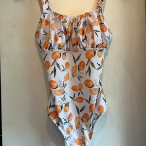 Vit baddräkt med apelsinmönster - Superfin vit baddräkt med färgglatt apelsinmönster och gröna blad. Axelband och lätt rynkad detalj vid bysten. Tillverkad i polyester och elastan för skön passform. Perfekt för strand eller pool! Endast testad! 