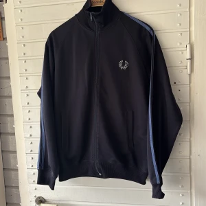 Marinblå track jacket från Fred Perry - Säljer en marinblå track jacket från Fred Perry med blå ränder längs ärmarna och broderad logga på bröstet. Jackan har dragkedja framtill, ribbade muddar och hög krage. Perfekt för en sportig och stilren look.