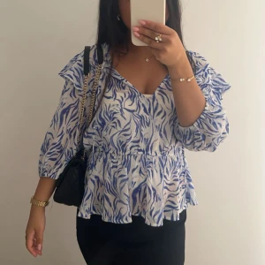 Blåvit blus med volang och v-ringning - Säljer en luftig blus med blått och vitt mönster, v-ringning och volangdetaljer vid axlar och nederkant. Blusen har trekvartsärm och är perfekt för vår och sommar. Supersnygg till både jeans och kjol!