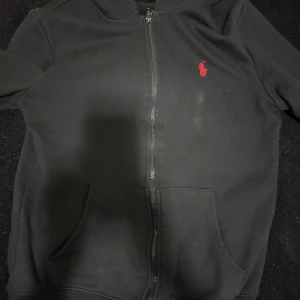 Svart hoodie från Polo Ralph Lauren - Svart hoodie från Polo Ralph Lauren med dragkedja framtill och röd broderad logga på bröstet. Tröjan har huva och två fickor framtill. Perfekt för en avslappnad och stilren look. OBS det är inte originala kedjan för kedjan gick sönder och jag var tvungen att byta den.