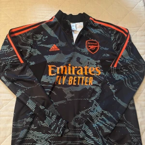 Arsenal långärmad träningströja från Adidas - Säljer en svart långärmad Arsenal träningströja från Adidas med orange detaljer och tryck. Tröjan har ett mönster i ljusblått och dragkedja vid halsen. Arsenal-logga och Emirates Fly Better-tryck på bröstet. 