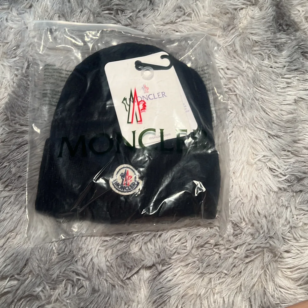 Svart ribbstickad mössa från Moncler med klassisk Moncler-logga framtill. Mössan är i ett mjukt material och har en enkel, stilren design som passar perfekt till kalla dagar.. Asusteet.