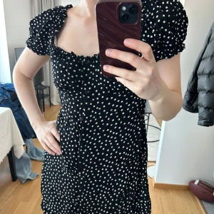Svart prickig klänning med puffärmar - Säljer en svart klänning med vita prickar och puffiga kortärmar. Klänningen har knappar framtill och en söt, lätt rynkad urringning. Figurnära passform och romantisk stil, perfekt för dig som gillar retrovibbar.