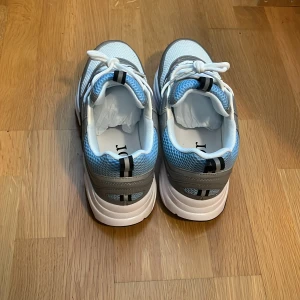 Grå Dior b22 - Bara seriösa köpare tack!!! Säljer ett par snygga Dior sneakers i grått, vitt och ljusblått med svarta detaljer. Skorna har tydlig Dior-logga på plösen och en chunky sula med blå inslag. Ovandelen är i mesh och syntetmaterial med vita skosnören. Med en digital kvitto med.