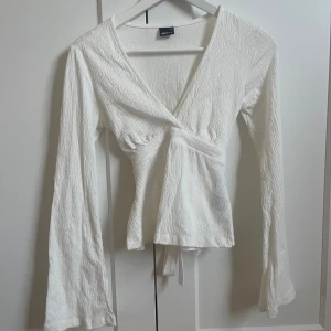 Vit topp med knyt från Gina Tricot - Säljer en vit topp från Gina Tricot i storlek XS. Toppen har lång ärm, v-ringning och knyt i midjan. Materialet är lätt strukturerat och stretchigt vilket ger en snygg passform. Perfekt till många olika outfits! Aldrig använt då det va ett litet hål på ena axeln när jag fick hem den💗