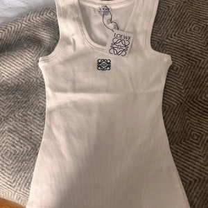 Vit ribbad linnetopp från Loewe - Säljer en stilren vit ribbad linnetopp från Loewe med broderad logga framtill. Toppen har en klassisk rund halsringning och är ärmlös, perfekt för en trendig och enkel look.