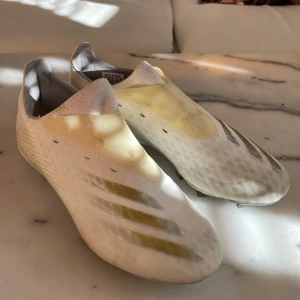 Vita fotbollsskor från Adidas - Snygga vita fotbollsskor från Adidas med en stilren design och diskreta gulddetaljer. Skorna har en strömlinjeformad form utan snörning för en smidig passform. Perfekta för fotbollsplanen med en modern look.
