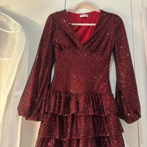 Vinröd paljettklänning med volanger från Meet Me There - Vinröd kortklänning med paljetter och v-ringning från Meet Me There. Klänningen har långa ärmar, flera lager volanger och glittrig yta. Perfekt för dig som vill sticka ut med en festlig look.