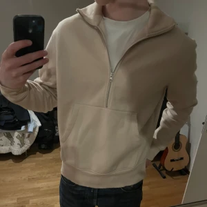 Beige halvzip hoodie från These Glory Days - Säljer en beige hoodie med halv dragkedja och stor magficka. Tröjan har hög krage och är tillverkad i mjukt material, perfekt för en avslappnad stil. Passformen är normal och ärmarna är långa.