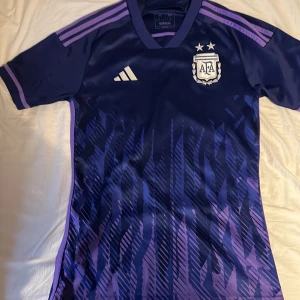 Argentina bortatröja 2022 från Adidas - Säljer en blå och lila Argentina bortatröja från Adidas i storlek XS. Tröjan har korta ärmar, mönstrad framsida och AFA-emblem med två stjärnor på bröstet. Klassiska Adidas-ränder på axlarna och tillverkad i ett lätt, syntetiskt material.