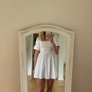 Vit klänning - Charmig vit kortklänning med puffärmar och smockad överdel. Perfekt för varma dagar med sin luftiga design och bekväma passform.